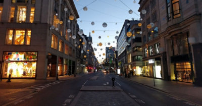 Prazna gradska ulica sa zgradama sa obe strane, osvetljenim izlozima i sfernim svetlosnim ukrasima koji vise iznad njih.