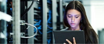 A woman in a server room is looking at a tablet. Server racks with cables are visible in the background.