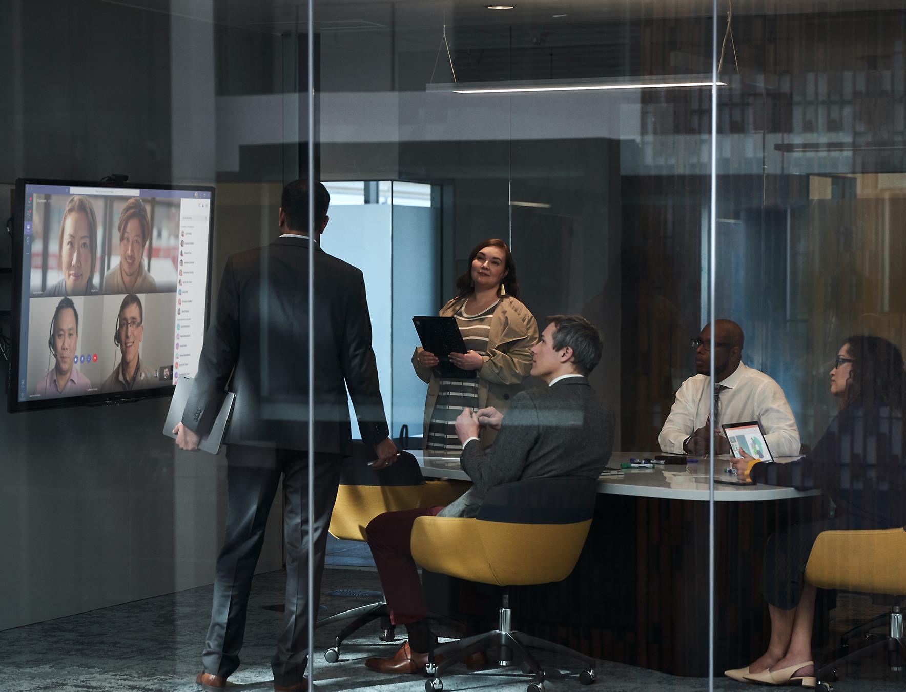 A group of people in a conference room meeting with people in other locations using Microsoft Teams displayed on a wall-mounted Surface Hub.