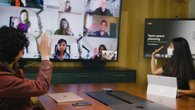 Two people wearing protective face masks sitting in a conference room participating in a Teams call being displayed on a television with many other participants.