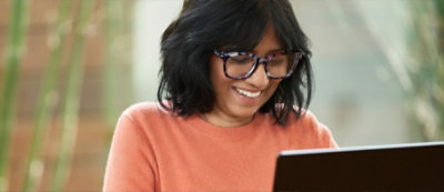 A person wearing glasses and an orange sweater smiles while looking at a laptop. 