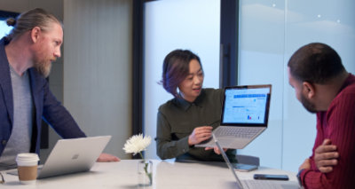 Three people working in a meeting room all with Surface laptops. One person is holding up their laptop and presenting the screen to the other two people.