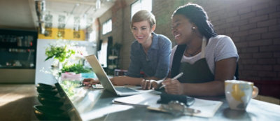 Dwie kobiety pracujące razem przy stole z laptopem, jedna uśmiechnięta, a druga trzymająca długopis, wewnątrz jasnego pokoju z roślinami i odsłoniętymi ceglanymi ścianami.