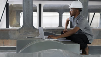 Person wearing a hard hat crouches down to use a laptop in a construction environment.