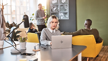 Laua taga istuv inimene töötab sülearvutiga ja kolm inimest peavad taustal mitteametlikku koosolekut