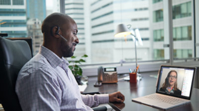 Person wearing a wireless earpiece sits at a desk and engages in a Teams meeting on a laptop.
