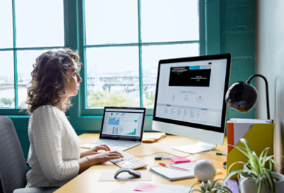 Foto di una persona seduta a una scrivania in ufficio, con un portatile e un grande monitor che digita