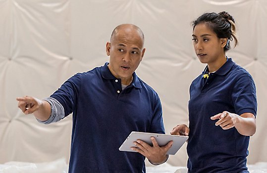 Photograph of two people standing in a high-end factory space talking and pointing while one holds a Surface in tablet mode