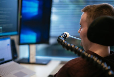 รูปถ่ายของผู้ทุพพลภาพกำลังใช้อุปกรณ์ Sip and Puff หรือ SNP ขณะนั่งอยู่บนรถเข็นที่โต๊ะทำงานที่มีแล็ปท็อปและจอแสดงผลหลายจอ