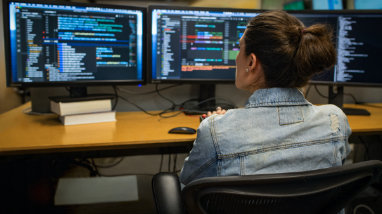 A person sitting at a desk looking at three monitors displaying information.