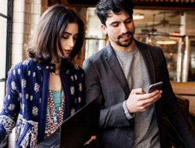 Two people having a conversation, one is using a mobile device, one is holding a laptop