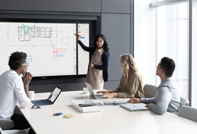 Three people in a conference room, with one standing and pointing at a large monitor displaying a diagram