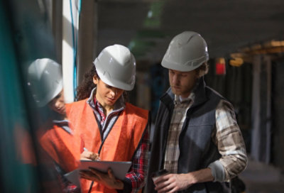 Two people standing in scientific facility wearing protective goggles and hair nets, looking at a device one of them is holding