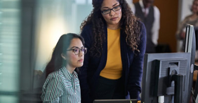 Фотографія двох людей, які дивляться на монітор настільного комп’ютера. Одна з них вводить текст за допомогою клавіатури, а інша показує на монітор.