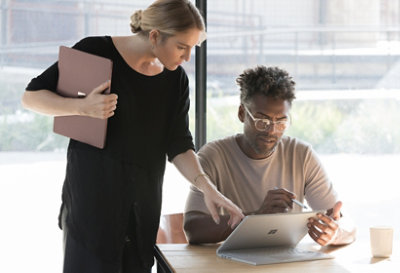 Deux personnes regardant l’écran d’un ordinateur portable Surface Book en mode studio. L’une d’elles est assise et tient un stylet, tandis que l’autre se tient debout et désigne l’écran tout en tenant un appareil Surface fermé.