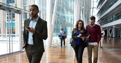 People with walking through a glass-enclosed atrium of a large modern building. One person is using a mobile device, another is using a tablet device.