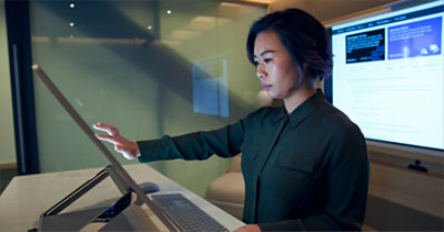 Eine Frau, die an einem Schreibtisch in einem Konferenzraum steht und einen großen Surface-Gerätemonitor berührt.