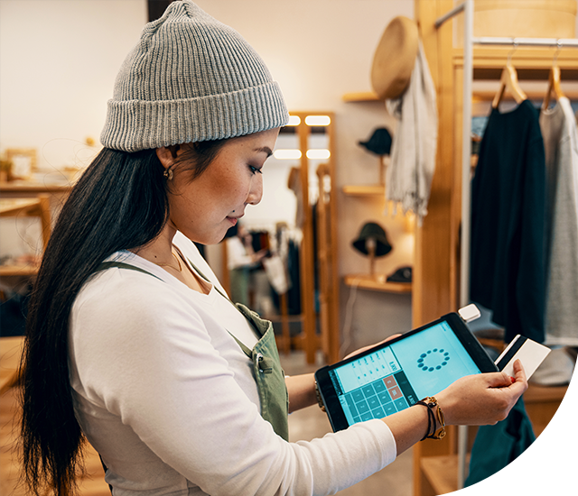 A person in a beanie and apron holds a tablet, swiping a card in a clothing store with hats and garments in the background.