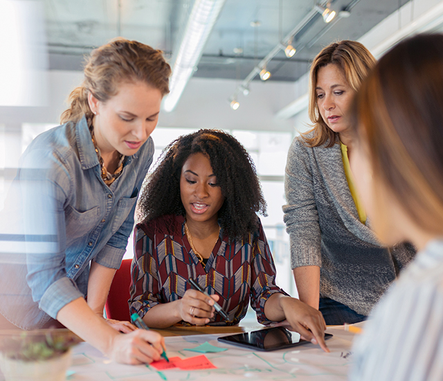 In a bright, modern office space, four women are engaged in a collaborative discussion around a table. The central figure, with curly hair, is writing on a large sheet covered with colorful sticky notes. The other women are attentively observing her work. They are all dressed casually, and the atmosphere conveys teamwork and creativity.