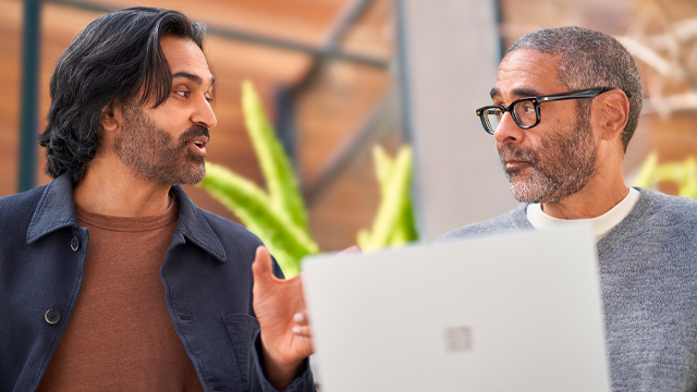 Two men are engaged in a conversation while looking at a laptop. The man on the left has long, dark hair and is animatedly speaking, while the man on the right, who has short hair and glasses, is listening thoughtfully. They are indoors, surrounded by greenery.