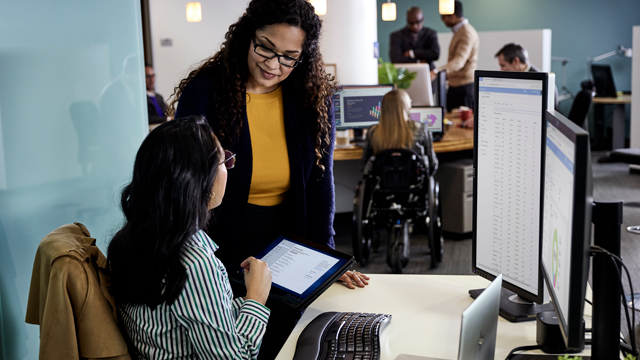 A woman in a yellow top and black pants stands beside another woman, who is sitting at a desk and looking at a tablet. The second woman wears a striped shirt and appears engaged in conversation. In the background, several colleagues are working at desks, including one individual in a wheelchair, highlighting a collaborative office environment.