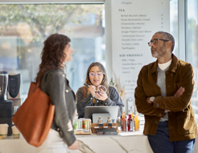Three people in conversation at a coffee shop counter; one person is a barista behind the counter