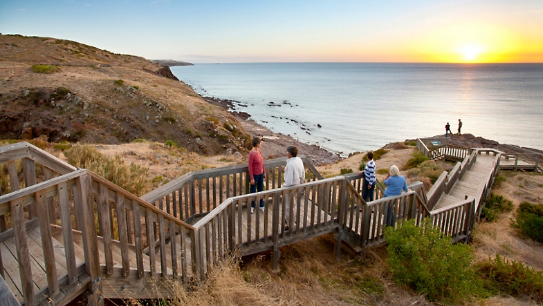People walking on a wooden boardwalk built into the hills along the ocean at sunset in southern Australia.