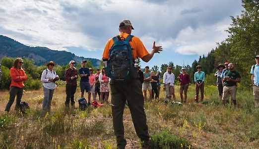 Un grupo de personas de pie escuchan a un guía en un campo con hierba.