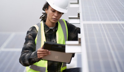 Eine Arbeiterin mit einer reflektierenden Sicherheitsweste und einem Schutzhelm, die ein Solarpanel untersucht und auf ein Tablet schaut.