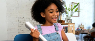 Child in a denim jumper holding a diploma in a room with a white brick wall and plants in the background.