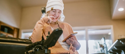 a woman in a white beanie and orange sweater sitting in a wheelchair and looking at her phone