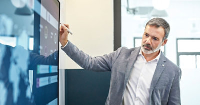 Muškarac sa kratkom sedom kosom i bradom, odeven u svetlosivi sako, koristi olovku za interakciju sa dodirnim ekranom 