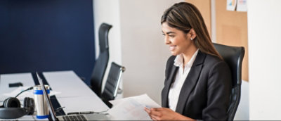 A professional woman in a black suit sitting at a desk with a document in hand, in an office setting.