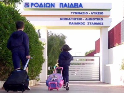 Zwei Kinder mit Rucksack-Trolleys gehen auf einen Schuleingang zu. Über diesem hängt ein Schriftzug in griechischer Schrift.