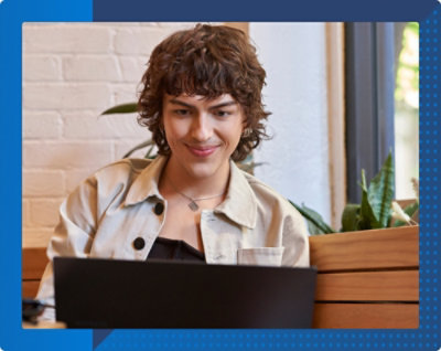 植物のある居心地の良い部屋でノート PCを見ながら微笑む巻き毛の人。