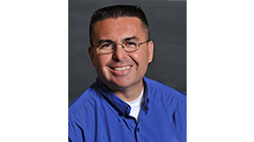 Smiling person wearing glasses and a blue collared shirt against a dark background.