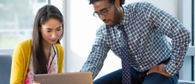 Two people with obscured faces, one seated with a laptop and the other standing, in a casual office setting.