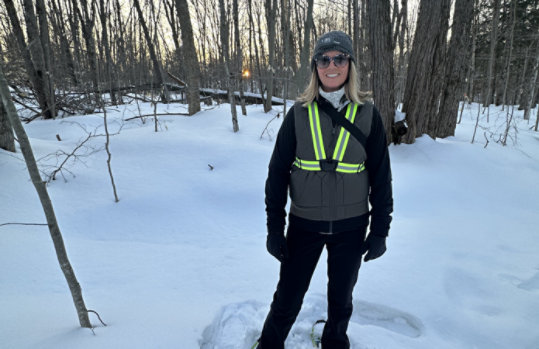  Karen Truyens clad in winter clothing and a reflective safety vest stands amidst a snowy forest, with bare trees