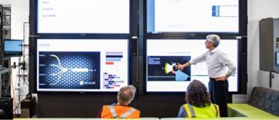 A presenter gesturing towards a graph on a screen during a meeting with two attendees in high-visibility vests.