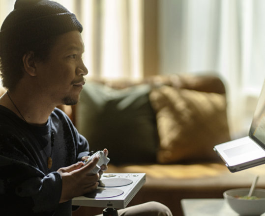 Xbox アダプティブ コントローラーとステージモードの Surface Laptop Studio 2 を使用するビーニー帽を被る若い男性。