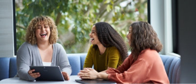 Three people sitting at a table, engaged in conversation and laughing, with a window and greenery in the background.