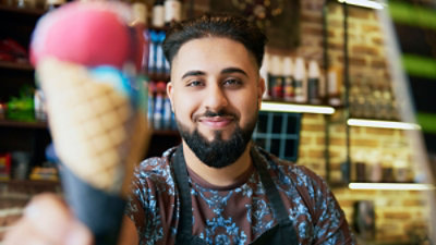 Muškarac u cvetnoj košulji i sa keceljom, osmehuje se u opremljenoj prodavnici i u ispruženoj ruci drži kornet sa dve kugle sladoleda.