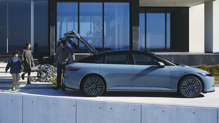 モダンなガラス張りの建物の前に停車したシルバーの電気自動車、リアハッチが開いていて人々がそばに立っている。