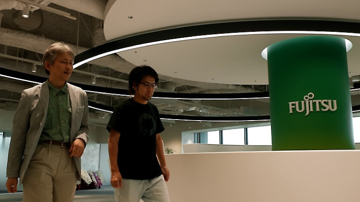 モダンなオフィス内を歩く二人と、円形の天井デザインに囲まれたFujitsuのロゴが表示されたグリーンの柱。