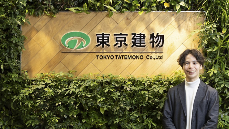 緑の植物に囲まれた東京建物（TOKYO TATEMONO Co., Ltd）の企業看板の前に立つグレーのブレザーを着た笑顔の男性。