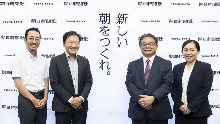 朝日新聞社のロゴと「新しい朝をつくれ。」のスローガンが書かれたバックパネルの前に立つ4人のビジネスパーソン。