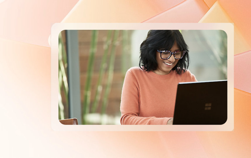 明るくモダンな空間で、背景に植物があるテーブルに座り、眼鏡をかけた笑顔の女性がノートパソコンで作業しています。