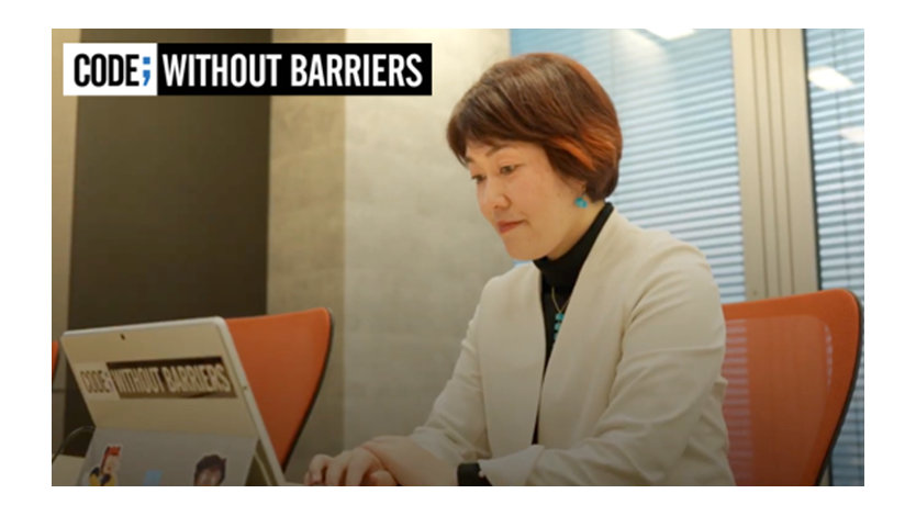 CODE; Without Barriers | 茶色のショートヘアの女性が机に座ってノートパソコンで作業しています。ノートパソコンには「Code; Without Barriers」と書かれたステッカーが貼られています。
