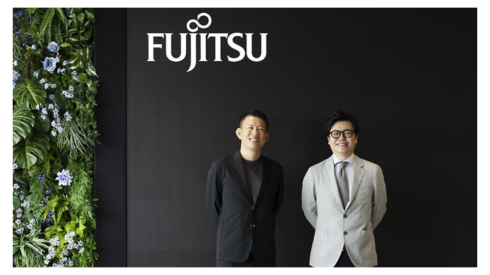 暗い壁のFUJITSUロゴと植物の壁の前に並んで立つ、スーツ姿の2人