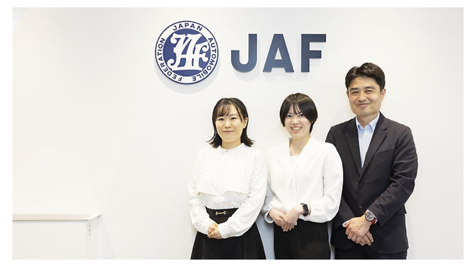 JAF（Japan Automobile Federation）のロゴと「JAF」の文字が掲げられたオフィスの壁の前に、3名が立っている様子。